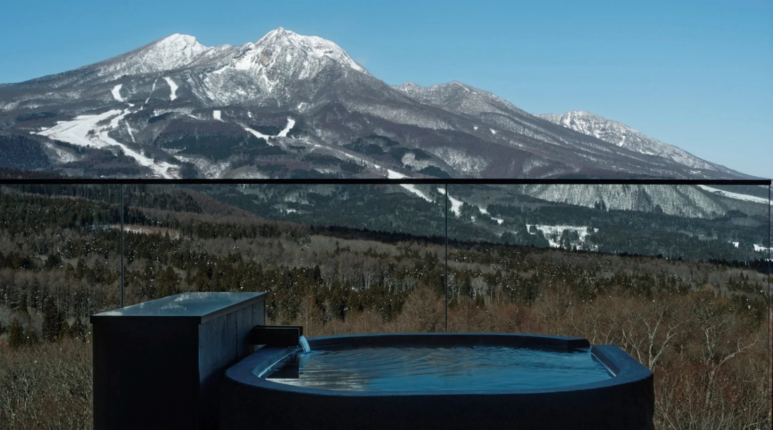 雪化粧した雄大な山々を背景に、広がる森と一体化したような露天風呂の絶景。冬の澄んだ青空の下、温かい湯に浸かりながら最高の癒しを体験できる様子がうかがえます。