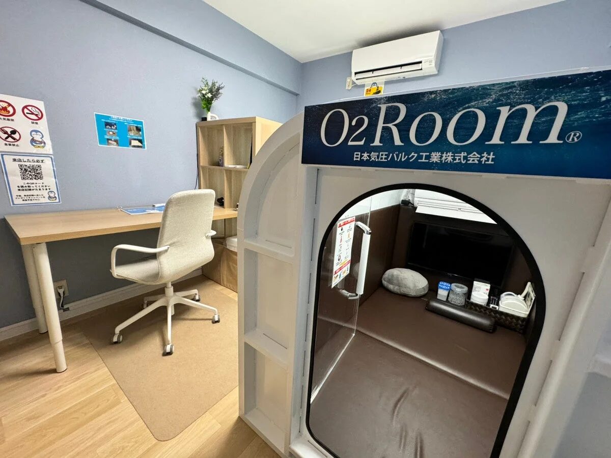 水色の壁の部屋に設置された酸素カプセル「O2Room」と、その手前にある受付用のデスクと椅子が写っています。