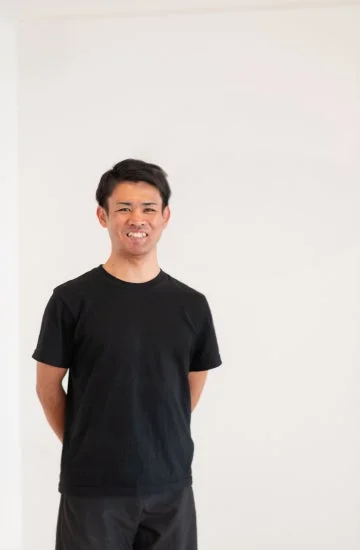 白い背景の前で黒いTシャツとパンツを着た男性が、カメラに向かって笑顔で立っているポートレート写真