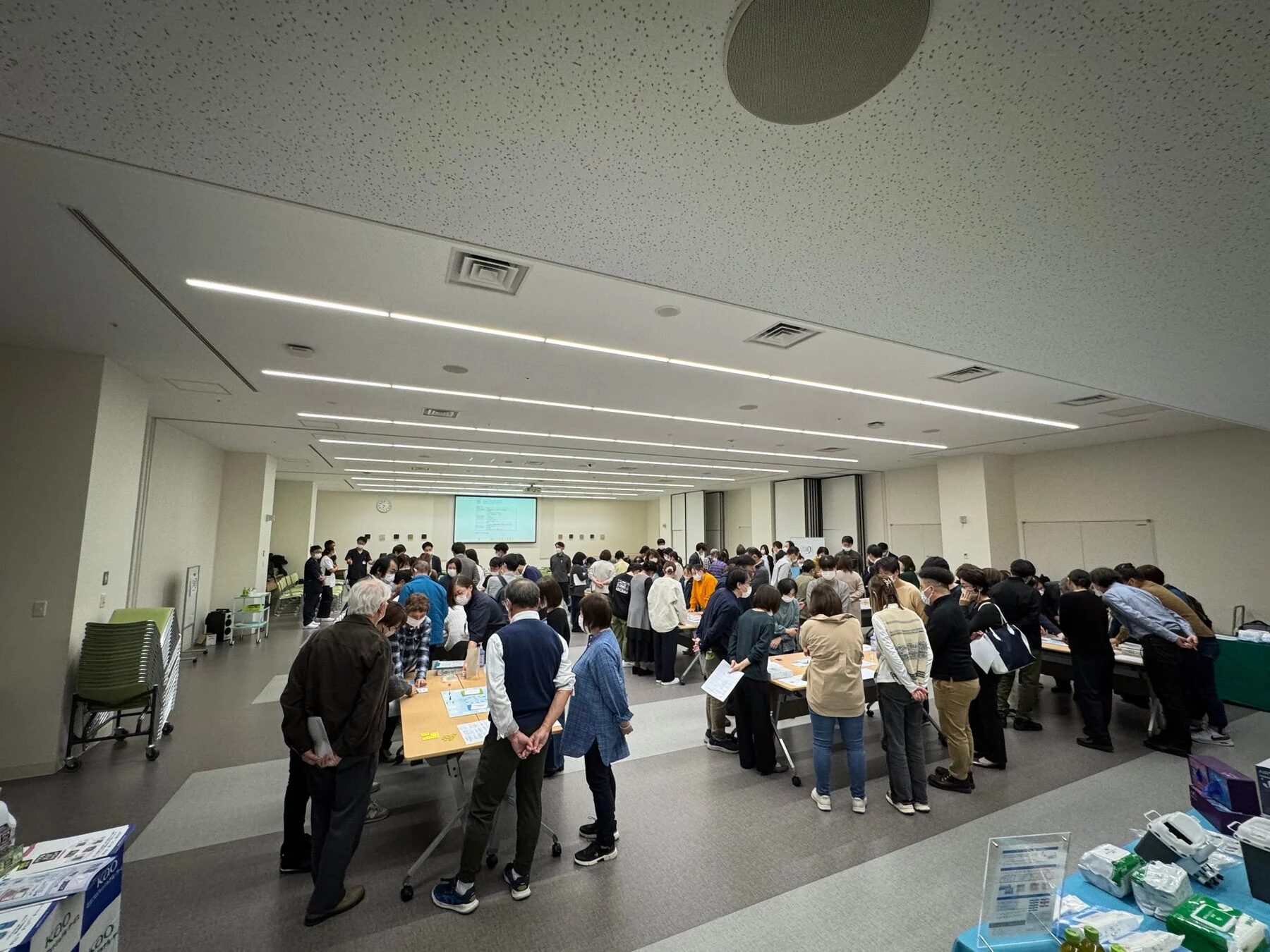 広い会場に多くの人々が集まり、テーブルを囲んで展示物や資料を見たり、互いに交流したりしている様子