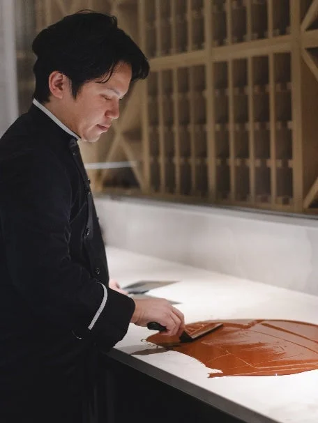 黒いコック服を着た男性が、白い作業台の上でチョコレートのような茶色い素材をスクレーパーで均している様子。真剣な表情で菓子作りに取り組む職人の手作業を捉えた一枚です。