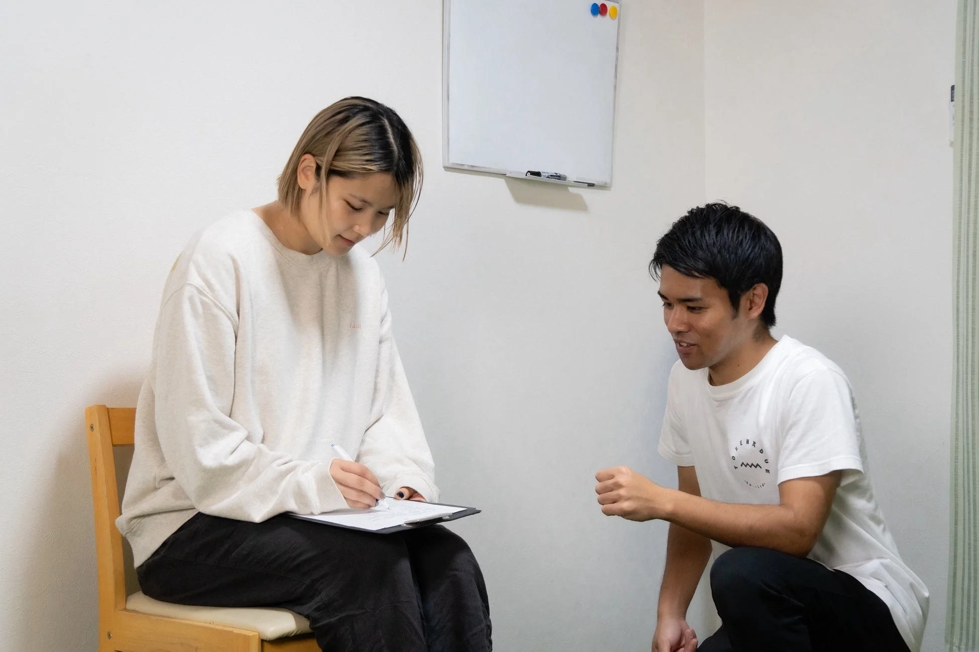 女性が椅子に座ってクリップボードに書き込み、男性がその前にしゃがんで話している様子