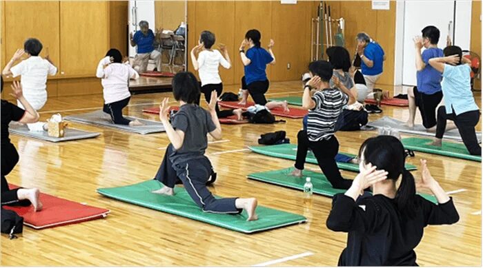 体育館のような場所で、複数の人々がヨガマットを使ってストレッチや体操を行っています。健康増進のためのグループエクササイズ風景です。