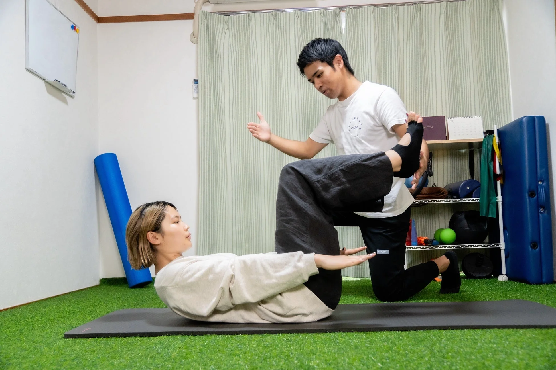 女性がヨガマットの上で脚上げ運動をしており、男性トレーナーが指導・補助している様子