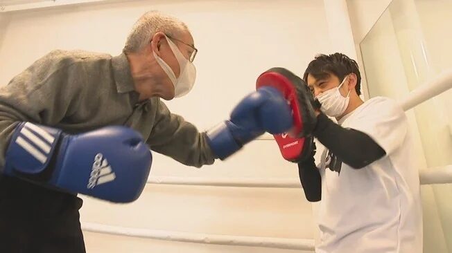 マスクを着用した高齢の男性が、ボクシンググローブを着けて、若いトレーナーが持つミットにパンチを繰り出している。ボクシングジムでのフィットネス風景で、高齢者の健康的な活動を象徴している。