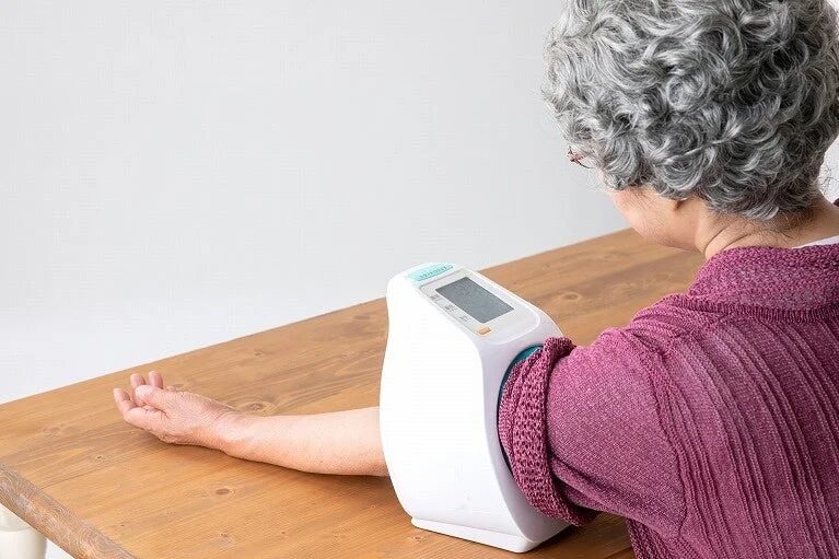 木製のテーブルに座り、腕に装着するタイプの血圧計で血圧を測定している高齢女性の後ろ姿です。日々の健康管理を行っている様子がうかがえます。
