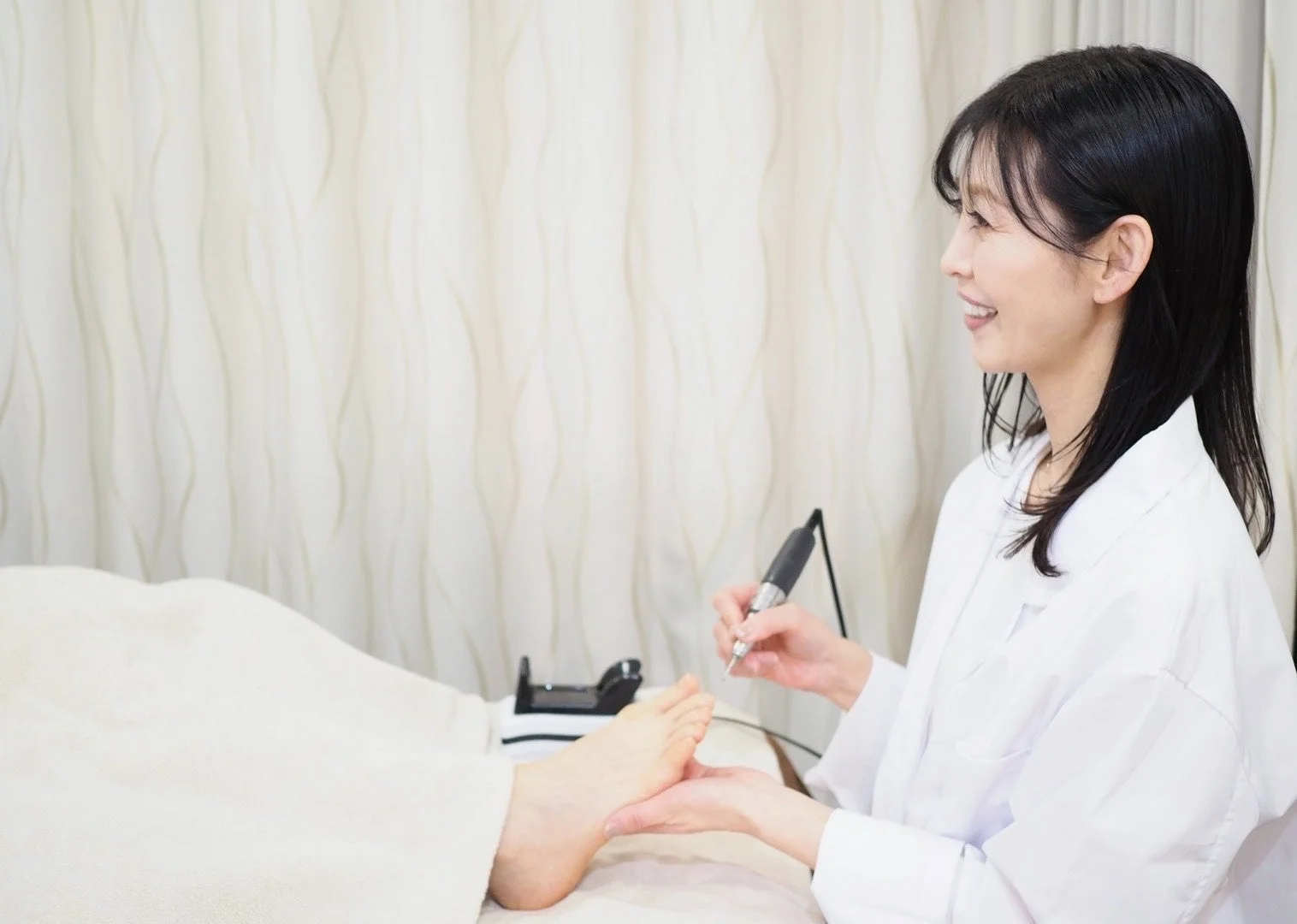 白衣を着た女性専門家が、フットケア用の電動ツールを使って顧客の足に施術を施している様子です。