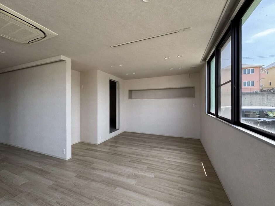 採光の良い広々とした空室の部屋。天井にはエアコンとダウンライトがあり、木目調のフローリングと白い壁が特徴的です。窓からは外の景色が見えます。