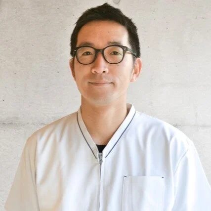 眼鏡をかけたアジア人男性が白いユニフォームを着用し、カメラに向かって穏やかな笑顔を見せている上半身のポートレート。