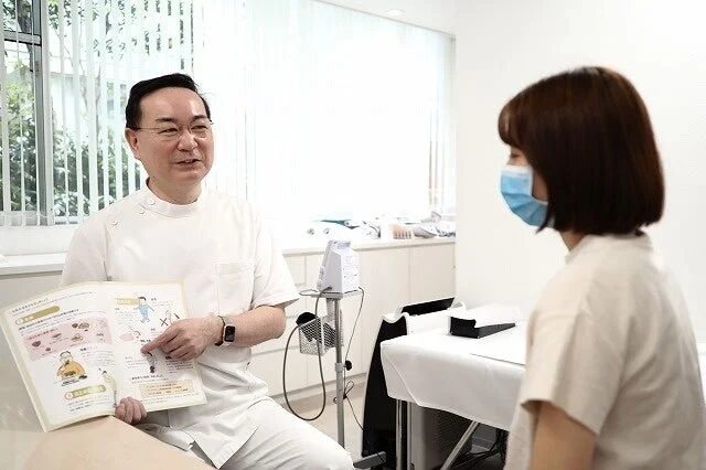 クリニックで医師が患者に対して、姿勢や生活習慣に関するイラスト入りの資料を見せながら丁寧に説明を行っています。患者はマスクを着用し、真剣に話を聞いています。