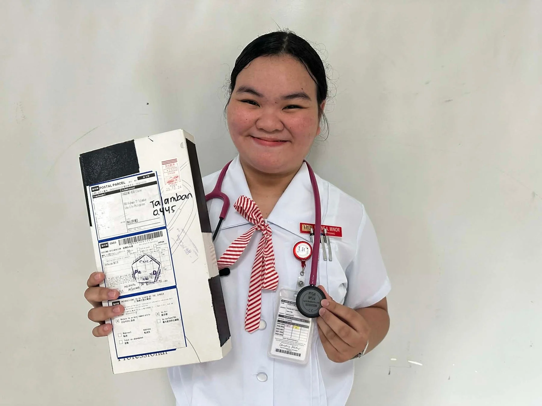 看護師か看護学生と思われる制服姿の若い女性が笑顔で小包と聴診器を持っている写真です。小包には宛先情報が記載されています。
