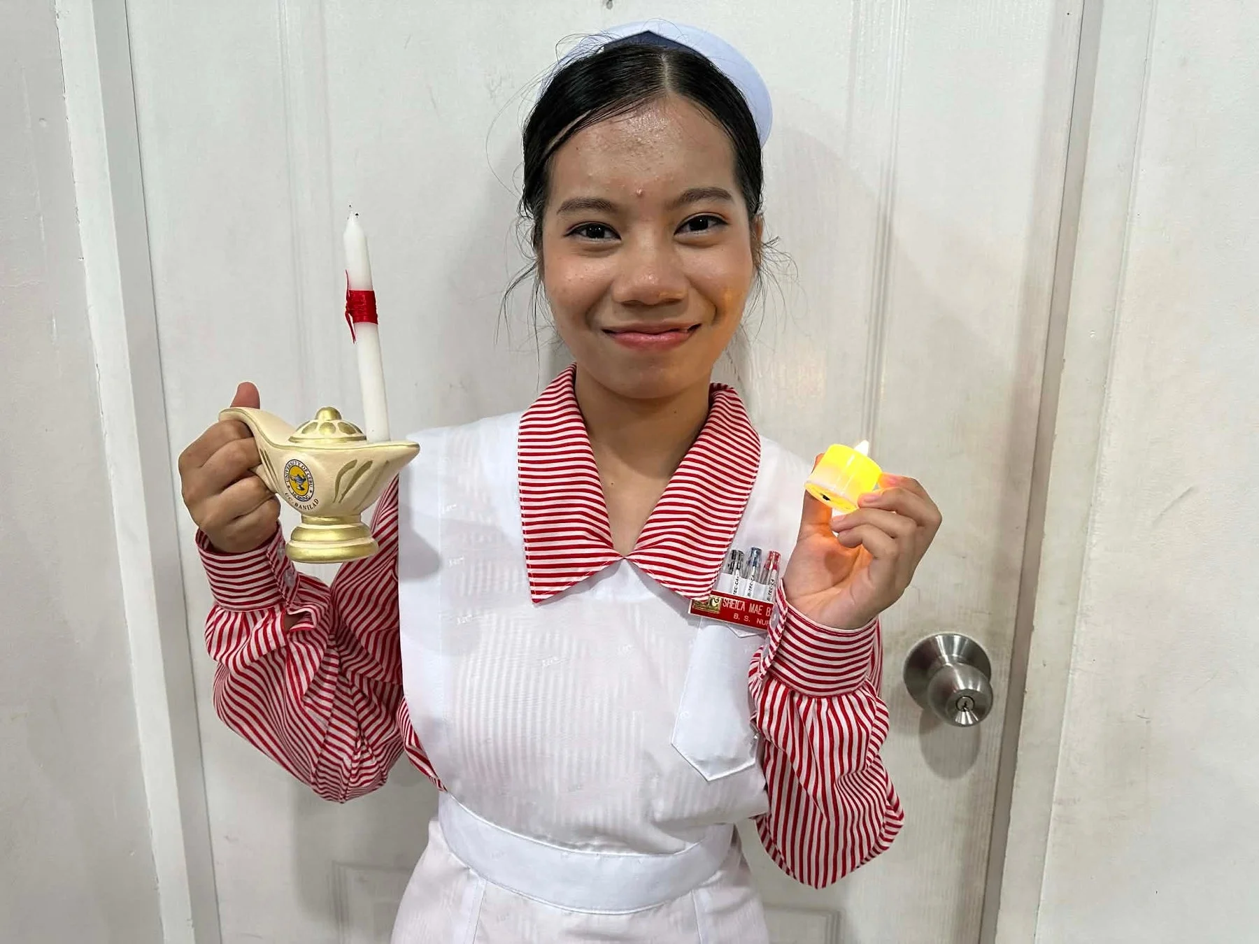 看護師の制服を着た女性が、ナイチンゲールランプとLEDキャンドルを持って笑顔で立っています。バッジには名前と「B. S. NURSING」と書かれており、看護学生のようです。