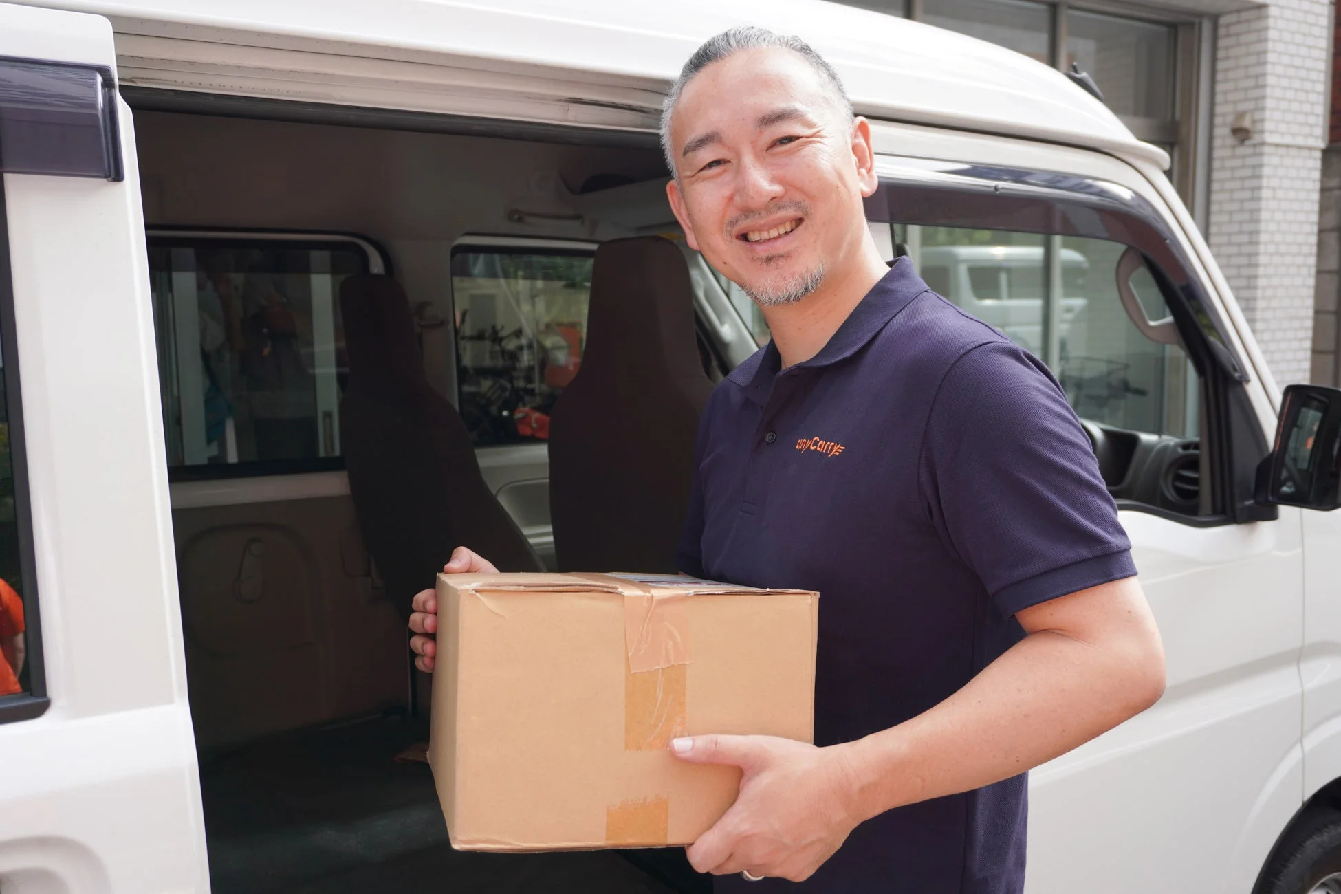 紺色のポロシャツを着た笑顔の男性が、白い軽バンの横で段ボール箱を抱えています。配送中のドライバーが荷物を運んでいる様子です。