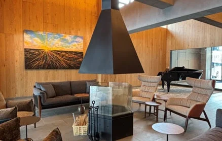 木製の壁が特徴的なモダンで広々としたラウンジの様子。中央には印象的な黒い暖炉があり、快適なソファやアームチェア、グランドピアノ、そして壁には抽象画が飾られています。