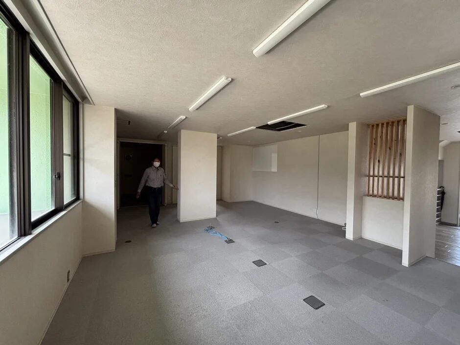 マスクを着用した男性が広々とした空のオフィススペースを歩いている様子。大きな窓とグレーのカーペットが特徴的で、内見や改装前の状態を思わせる空間です。