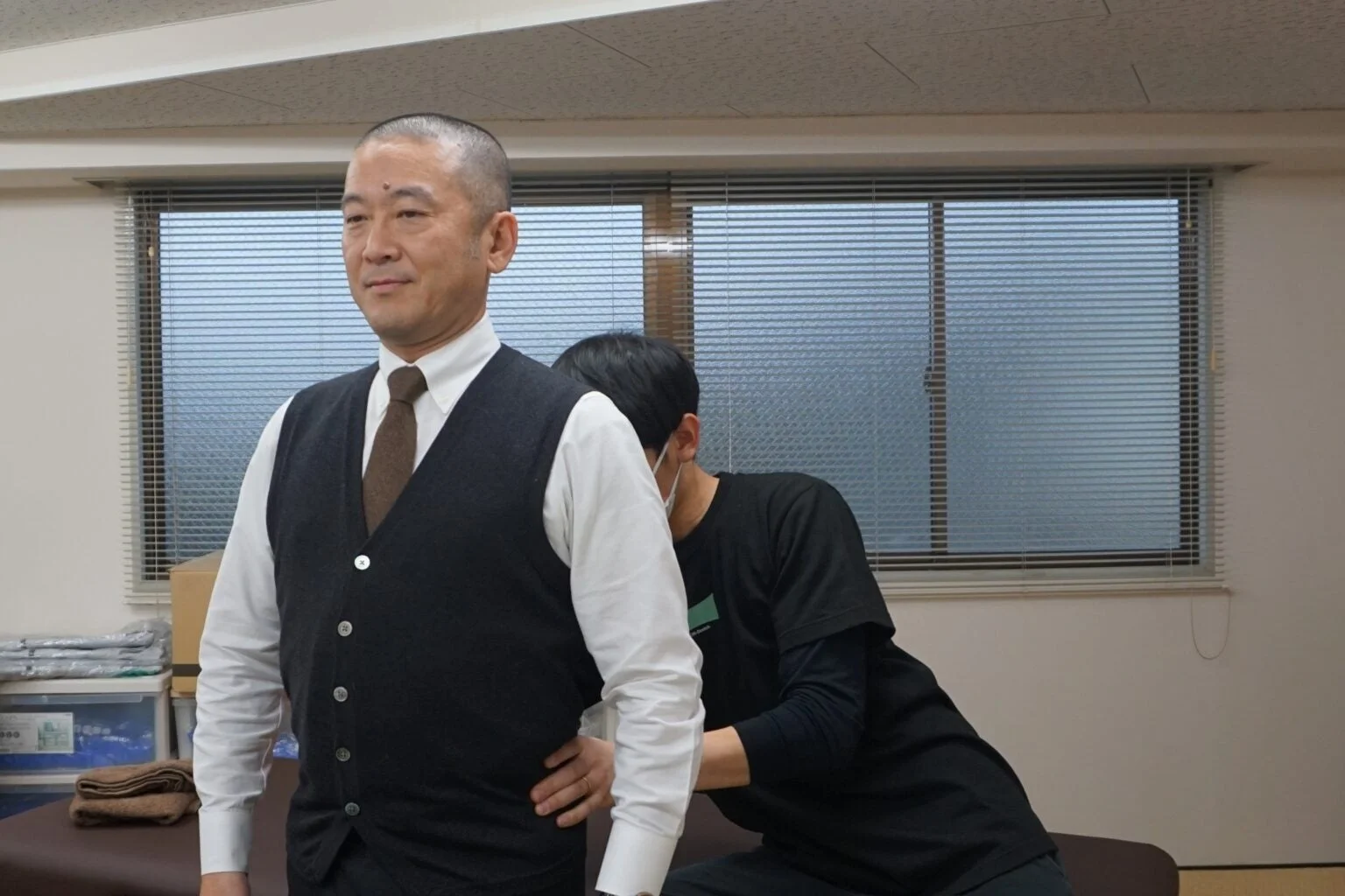 室内で、スーツを着た男性が黒いシャツを着た施術者から腰の施術を受けている様子。整体院や治療院でのケアを思わせる。