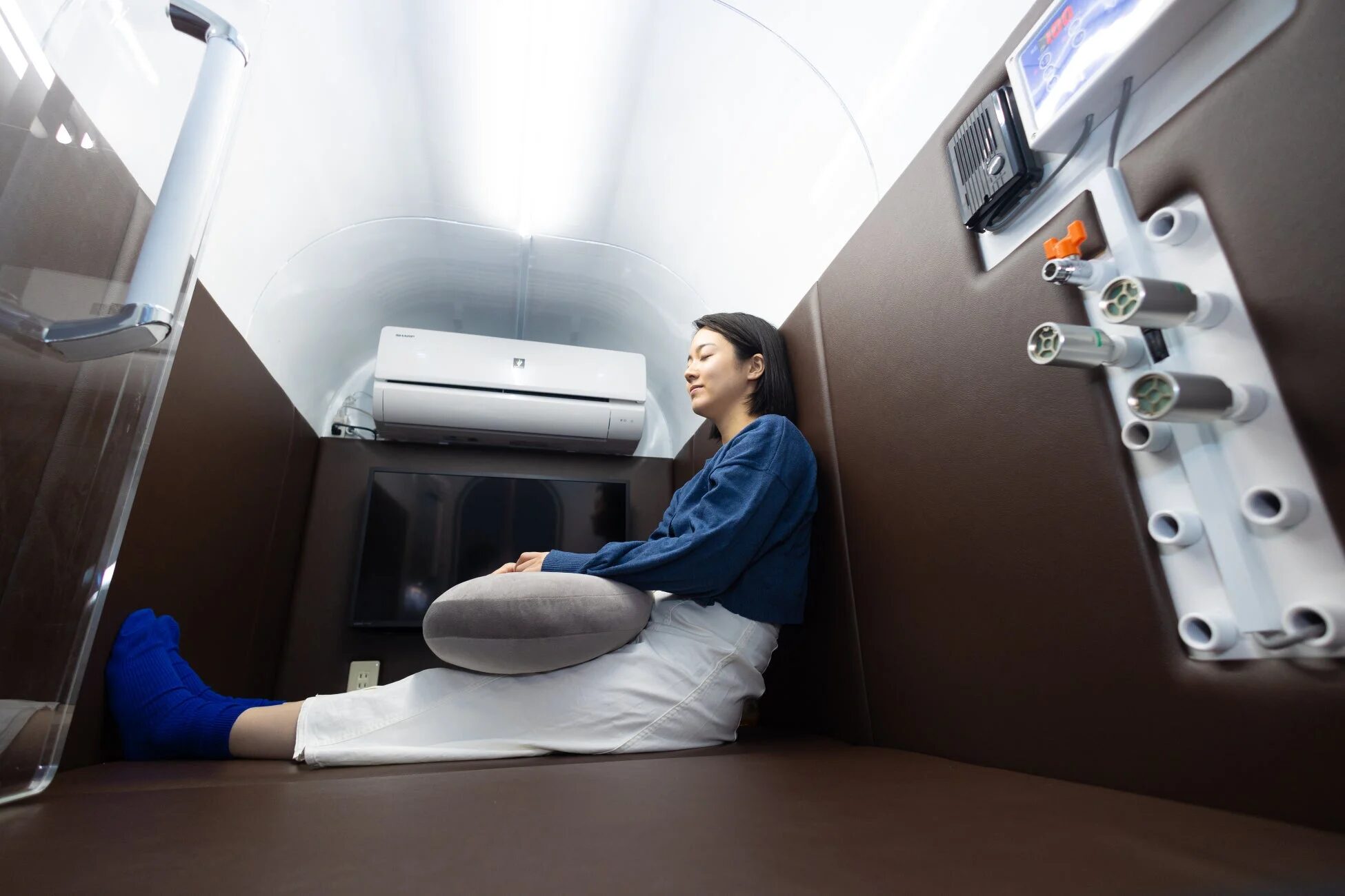 現代的なカプセル型個室で女性がリラックスしている様子。エアコンや操作パネルがあり、快適な休息スペースのようだ。