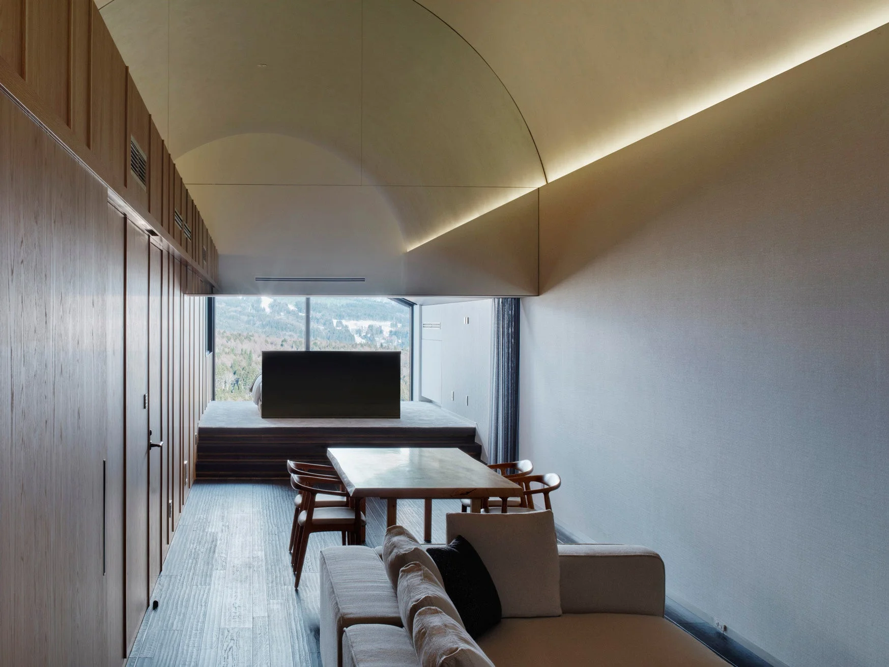 モダンで洗練されたリビングダイニングルームの風景。大きな窓からは雪に覆われた山々の景色が広がり、自然と調和した開放的な空間が特徴です。木材を基調とした内装と曲線的な天井の間接照明が落ち着いた雰囲気を演出しています。