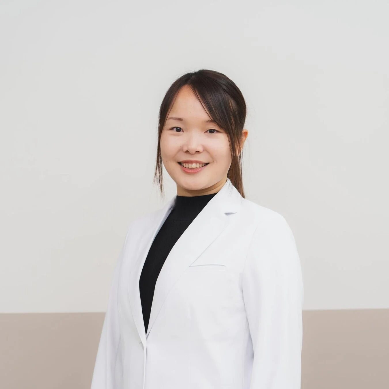 白衣を着て笑顔でカメラを見つめる若いアジア人女性のポートレート。清潔感のある背景で、専門的で親しみやすい印象を与えます。