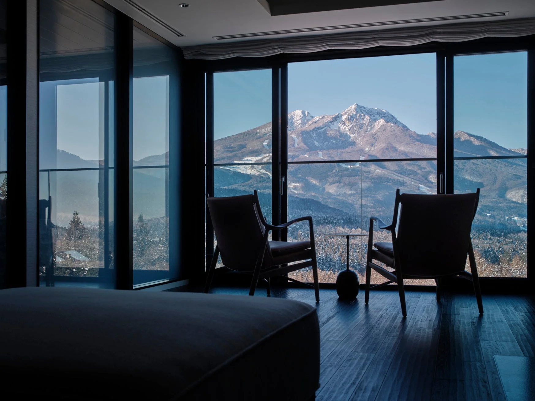窓から雪をまとった雄大な山々を望む、モダンで洗練された室内の様子。手前にはゆったりと座れる椅子が配置されており、絶景を楽しみながら贅沢な時間を過ごせる空間であることが伺えます。