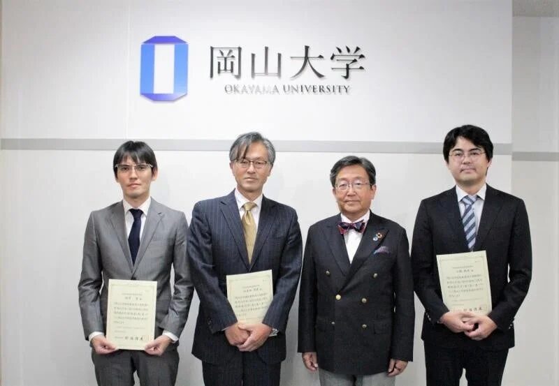 岡山大学のロゴの前で、スーツを着た4人の男性が賞状のような書類を持って並んでいる写真