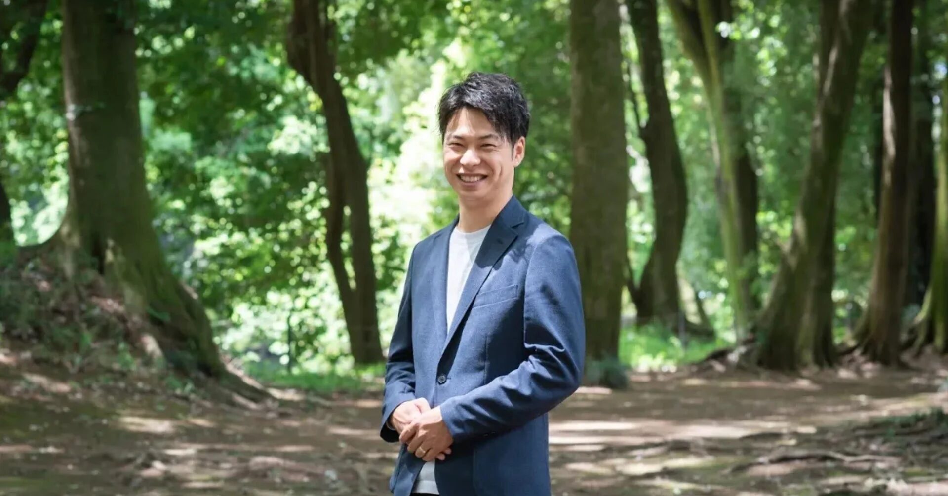 紺色のジャケットを着た男性が緑豊かな木々に囲まれた屋外で笑顔を見せている