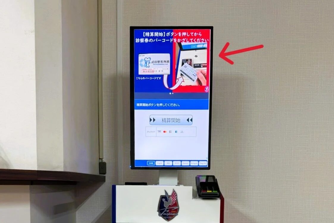 医療機関に設置された自動精算機が写っています。画面には診察券のバーコードをかざして精算を開始する手順が日本語で表示されており、複数の言語選択肢も見られます。