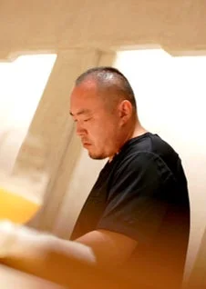 黒いTシャツを着た中年の男性が、真剣な表情でうつむいている横顔のショットです。短髪とひげが特徴的で、何か作業に集中している様子がうかがえます。