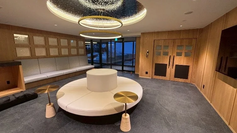 モダンで落ち着いた雰囲気のラウンジまたはロビー。木目調の壁と豪華な照明が特徴的で、長いソファと円形のベンチが配置された高級感のある洗練された空間です。