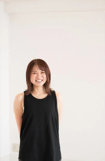 白い背景の前で、黒いタンクトップを着た若い女性が明るく笑顔を見せているポートレート写真