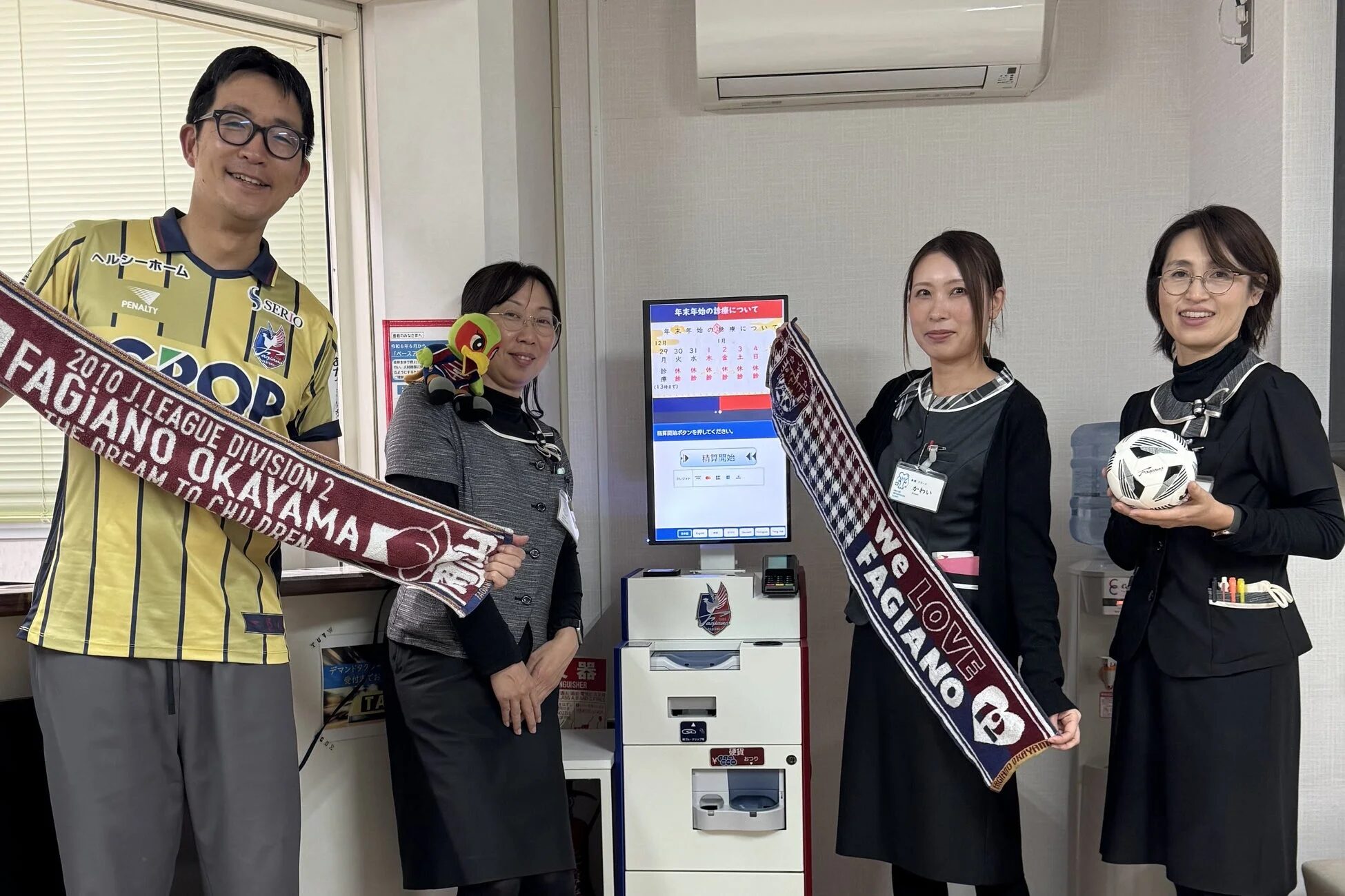 ファジアーノ岡山に関連するサッカーグッズを持った4人の男女が、クリニックのような施設で笑顔で立っている様子です。