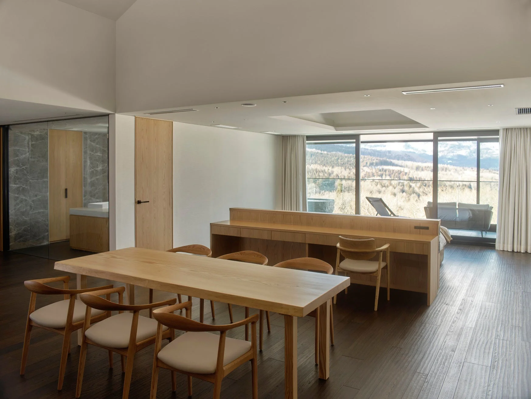 木製家具が配されたモダンで広々とした空間。大きな窓から雄大な山々を望む、自然光が差し込む落ち着いたリビング。奥には寝室やバスルームも見える、洗練されたリゾート風インテリアです。