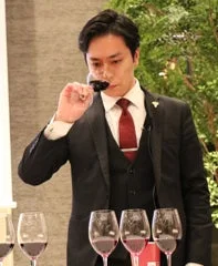 スーツを着た男性がワイングラスを鼻に近づけ、真剣な表情でワインのテイスティングをしている様子です。テーブルには複数のワイングラスが並べられており、専門的な試飲会やソムリエの仕事風景を思わせます。