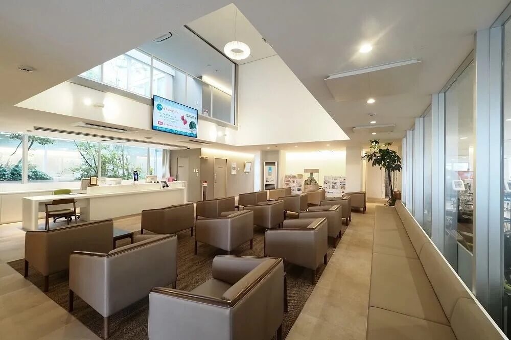 広々としたモダンで清潔感のあるクリニックの待合室です。受付カウンターと快適そうな椅子が並び、窓からは緑豊かな庭が見えます。壁のテレビにはインフルエンザワクチンの案内が表示されています。