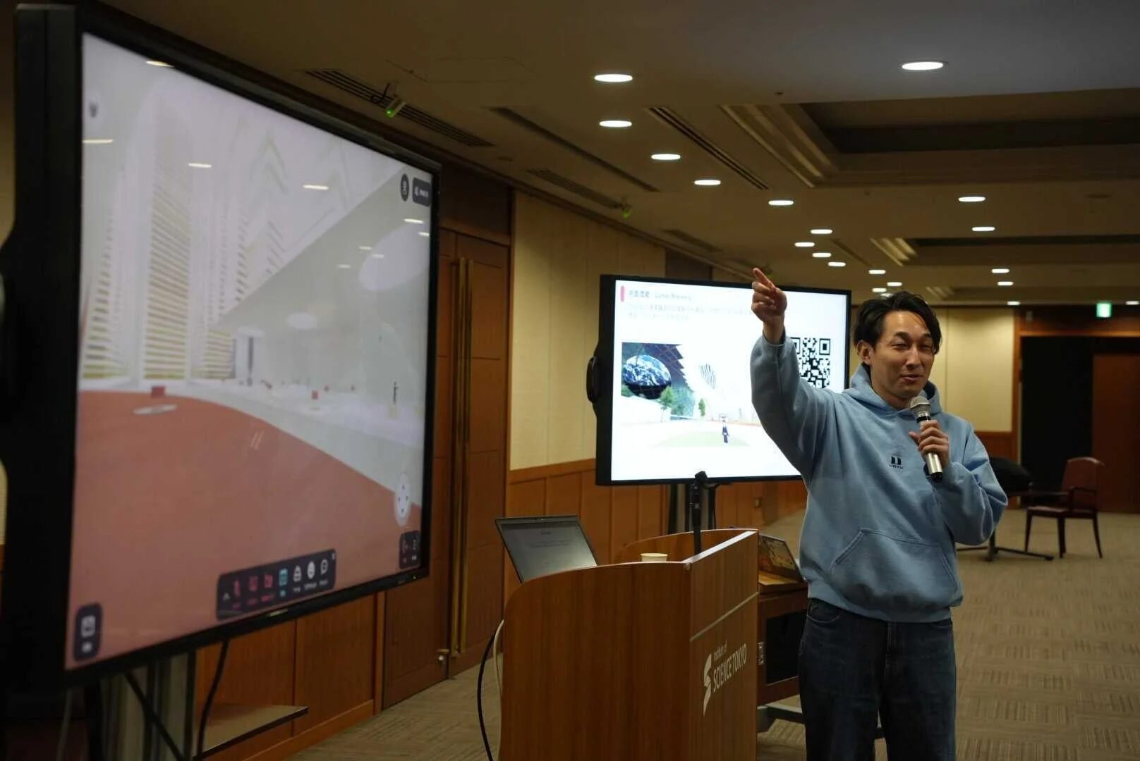 男性がマイクを手に、2つの大型スクリーンを背景にプレゼンテーションを行っています。