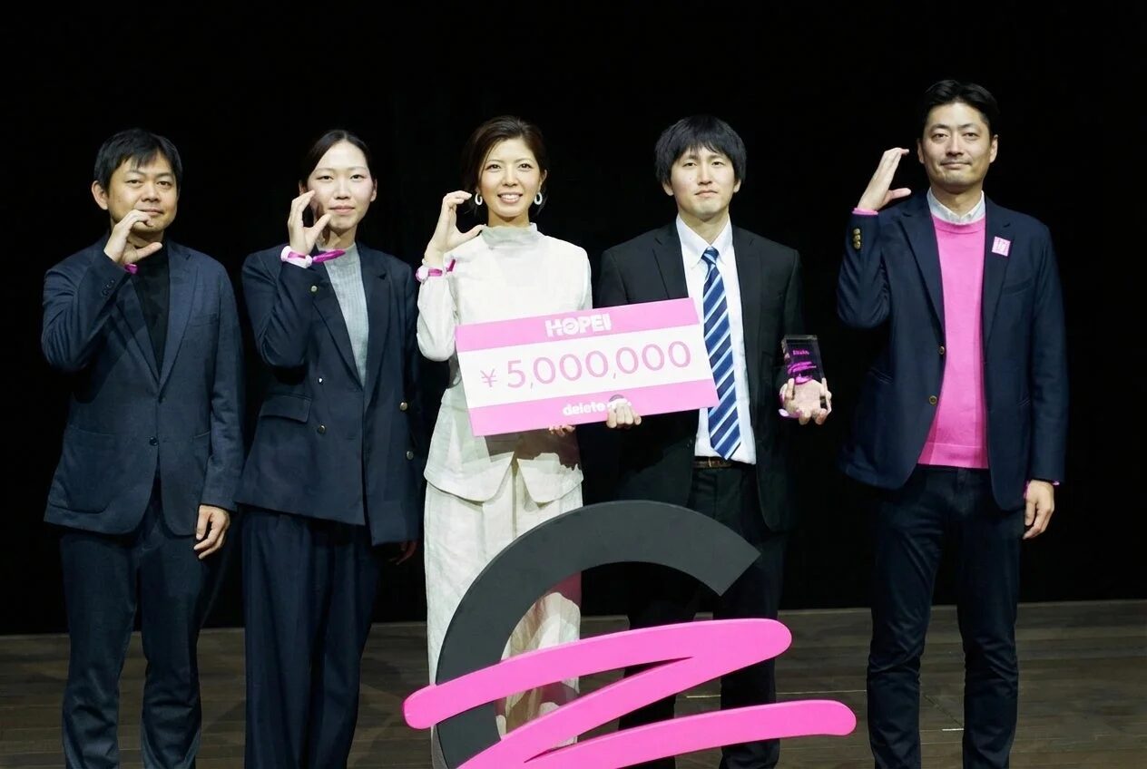 deleteCの授賞式で、500万円の賞金パネルとトロフィーを手に記念撮影に応じる牧野祐紀さんを含む5名の男女。喜びと成功が感じられる瞬間です。
