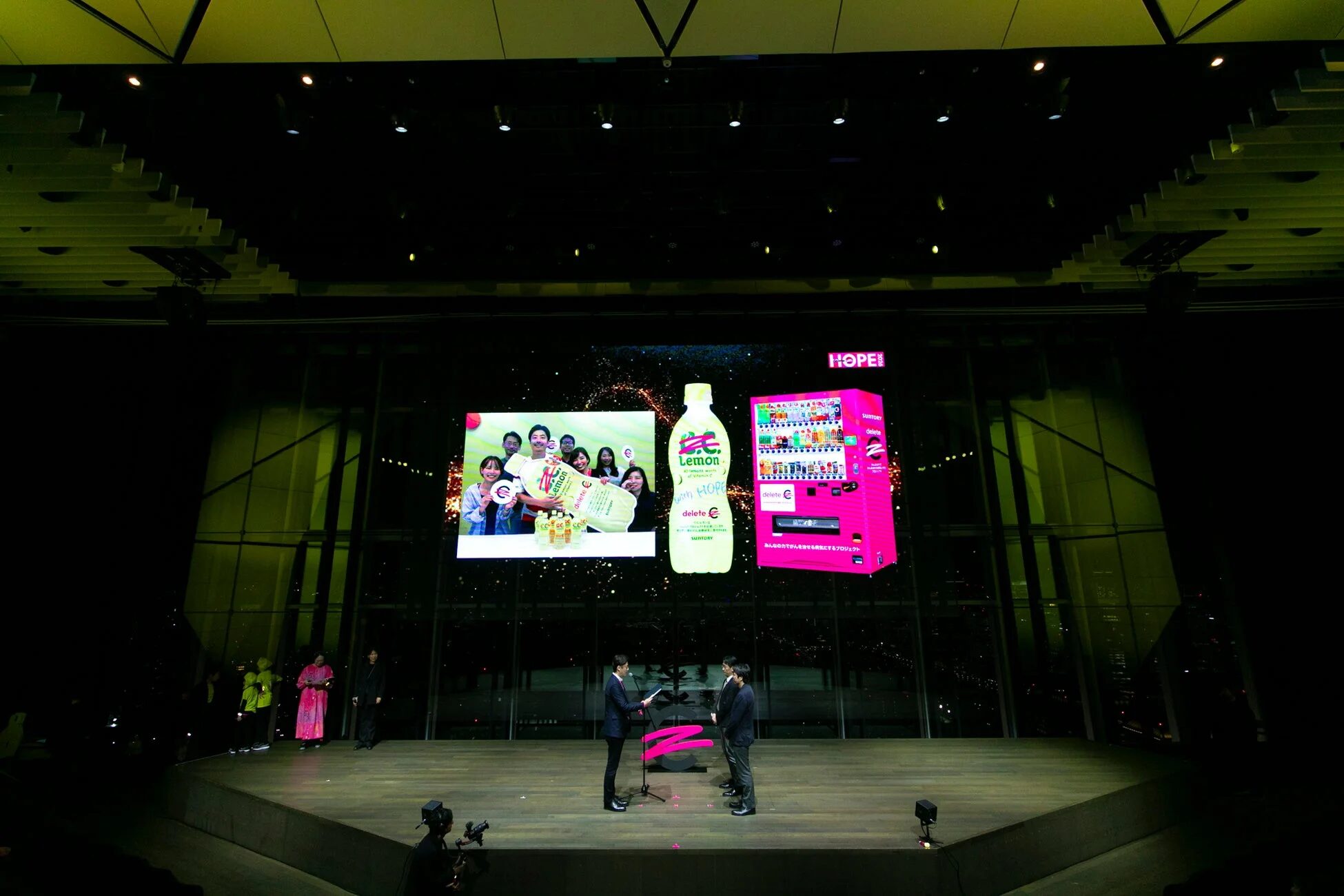 イベントのステージには、サントリーC.C.レモンやピンクの自動販売機が映し出された巨大スクリーンがあり、がん支援プロジェクトについて語られています。