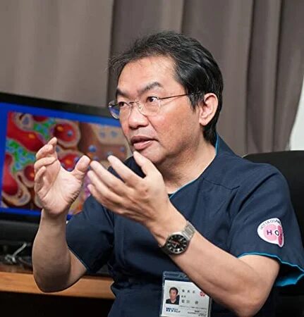 眼鏡をかけた中年の男性が、医療用スクラブを着て手を広げて話している。背景のモニターには、細胞らしきカラフルな画像が映し出されており、専門的な説明をしている様子がうかがえる。