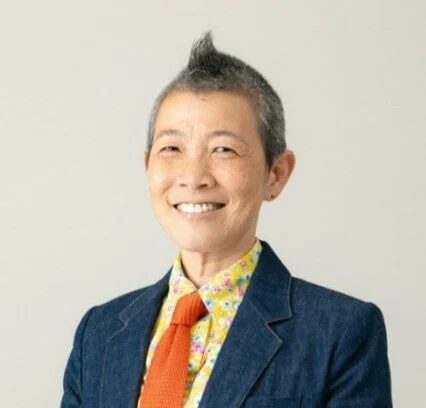 笑顔でカメラを見つめるアジア人女性のポートレート。短く白いモヒカン風の髪型で、デニムジャケットに花柄シャツとオレンジのネクタイを着用しています。明るい表情が印象的です。