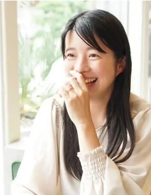 長髪の若い女性が、手を口元に当てながら明るく笑っているポートレート写真です。自然体で幸せそうな表情をしています。