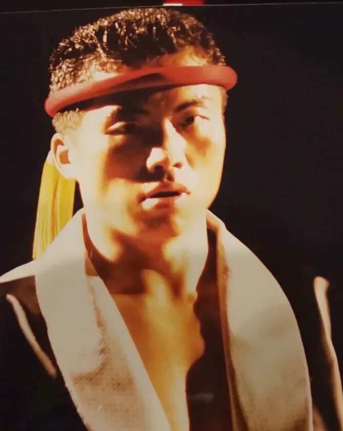 赤いヘッドバンドを着用した男性のポートレート。暗い背景の中で、顔には強い光が当たり、真剣な表情を浮かべている。格闘技に関連する雰囲気を持つ。