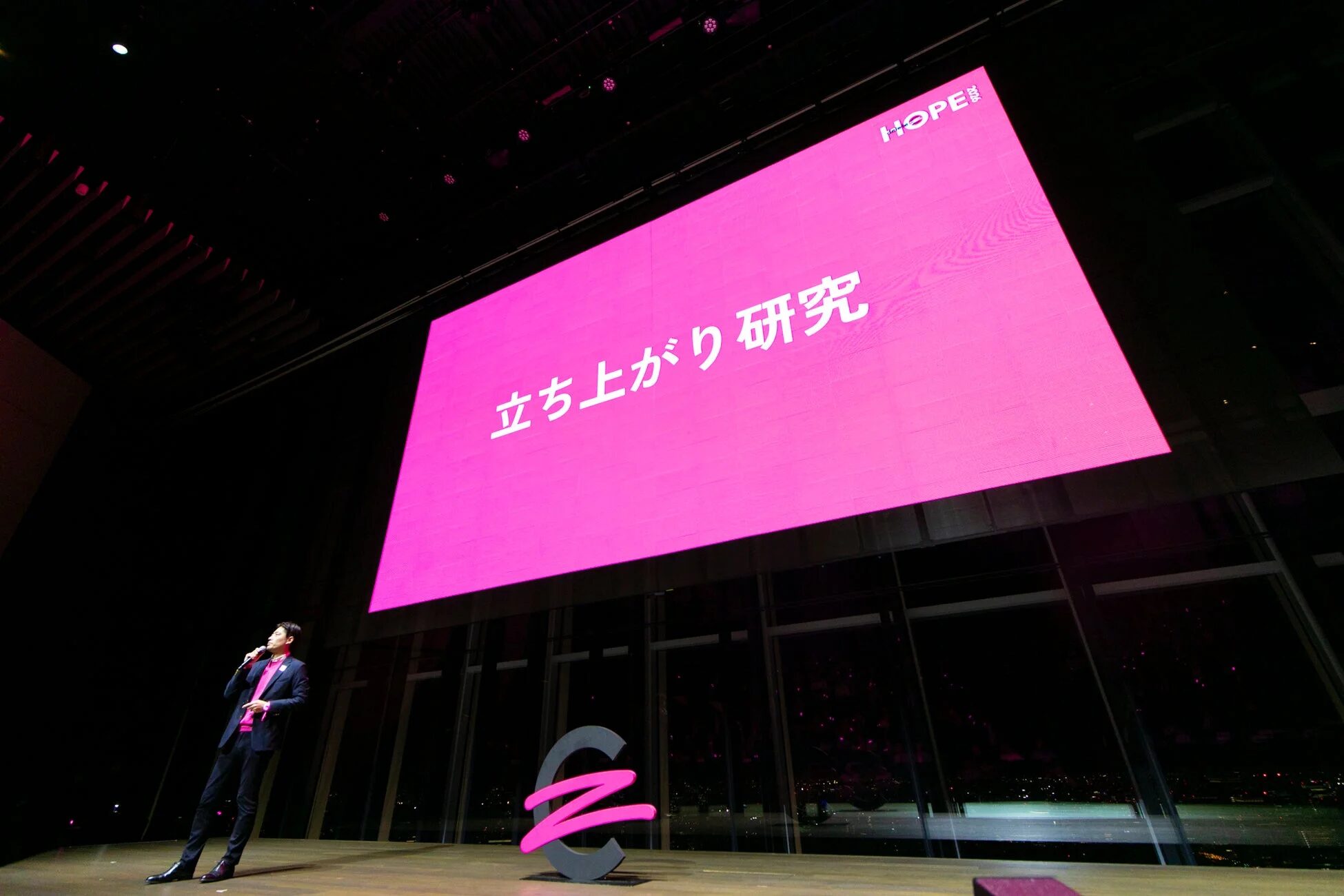 ステージ上でマイクを持って講演する男性。背後のスクリーンには「立ち上がり 研究」と「HOPE 2020」の文字が表示されています。