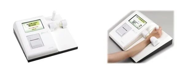白い医療用測定機器が2つのアングルで写されており、腕を置いて使用する様子が示されています。画面には氏名確認や患者情報が表示されており、健康診断や検査に使用される機器です。