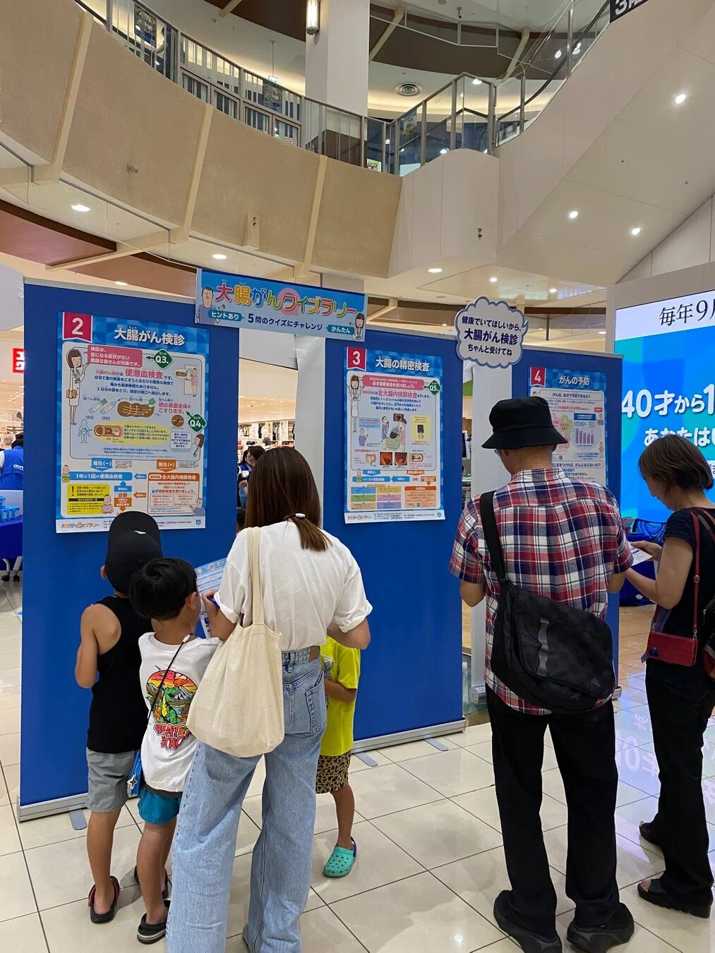 ショッピングモールで大腸がんの啓発イベントが開催されています。青いパネルに大腸がん検診や予防の情報が展示され、大人や子供たちがクイズを解きながら学んでいます。40歳からの検診が促されています。