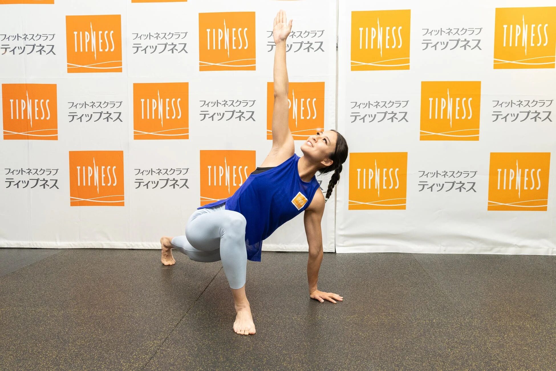 フィットネスクラブ「ティップネス」のロゴが背景に並ぶ空間で、スポーツウェアを着用した女性がヨガやストレッチのようなポーズをとっています。片腕を高く上げ、体をひねりながらバランスをとっており、健康的な運動の様子を捉えた一枚です。