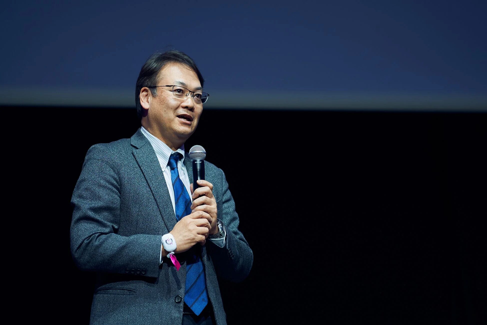 マイクを手にスピーチをするスーツ姿の男性。真剣な表情で、イベントでプレゼンテーションを行っています。