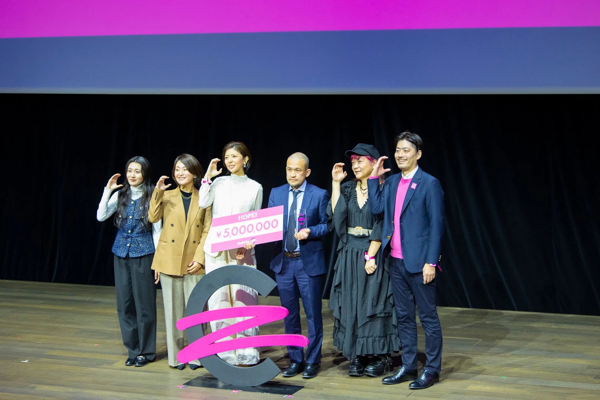 deleteCの授賞式で、500万円の賞金パネルとトロフィーを持って記念撮影をする児玉真さんを含む数名の男女。ステージ上での喜びの瞬間です。