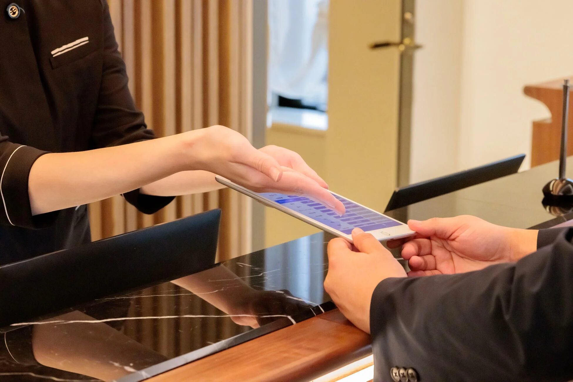 受付カウンターでスタッフが顧客の持つタブレットを操作し、情報入力や案内を行っている様子
