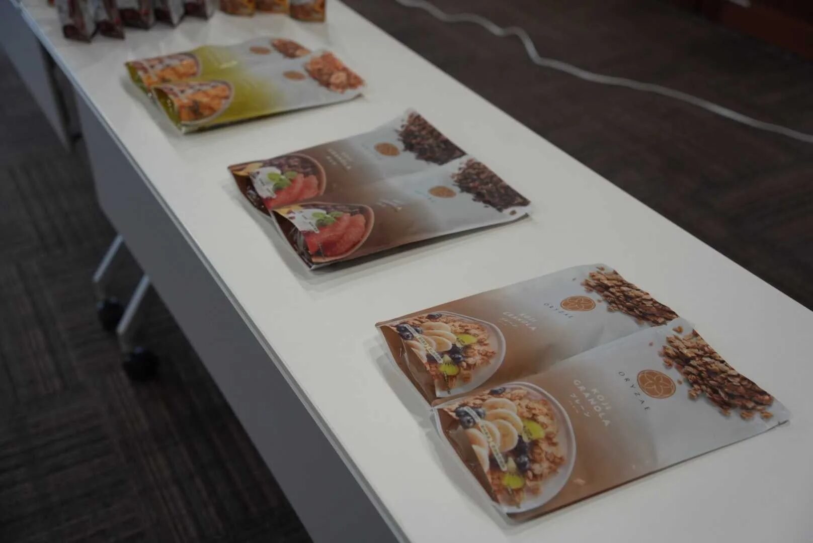 白いテーブルに「ORYZAE KOJI GRANOLA」のパッケージが複数陳列されています。