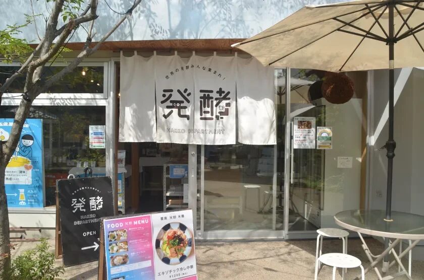 「発酵」をテーマにした飲食店「HAKKO DEPARTMENT」の外観です。白い暖簾とメニューボードが目を引き、テラス席にはパラソルが設置されています。